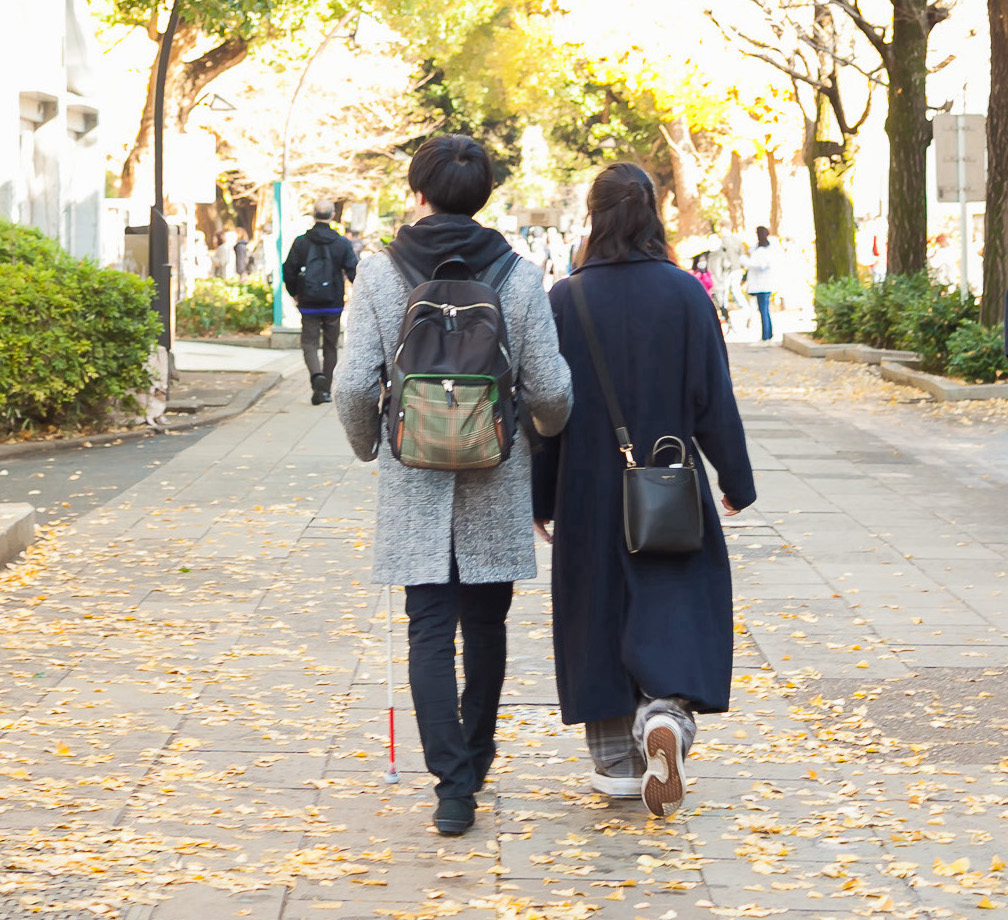 白杖を持った男性との女性ヘルパーが、街なかを歩いている様子
